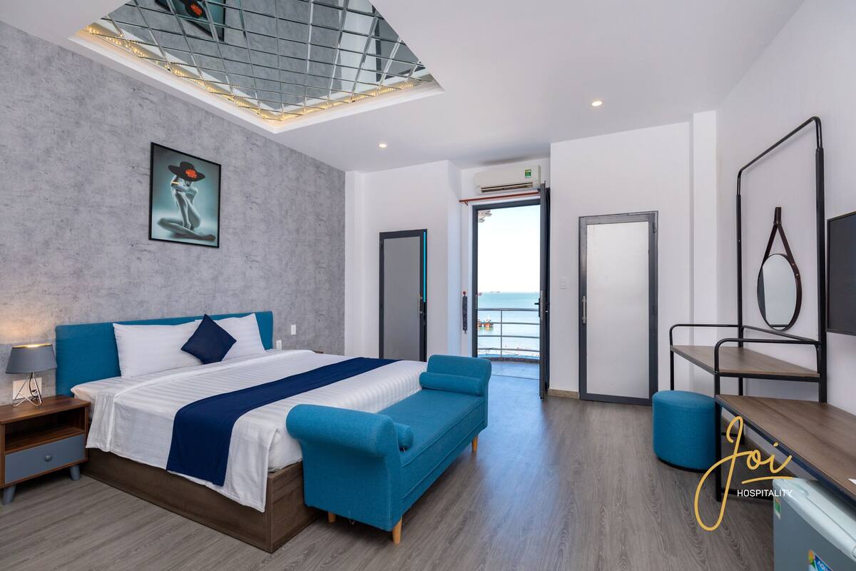 Habitación de hotel moderna con una cama grande, un sofá azul, un escritorio y vista al mar a través de la puerta.