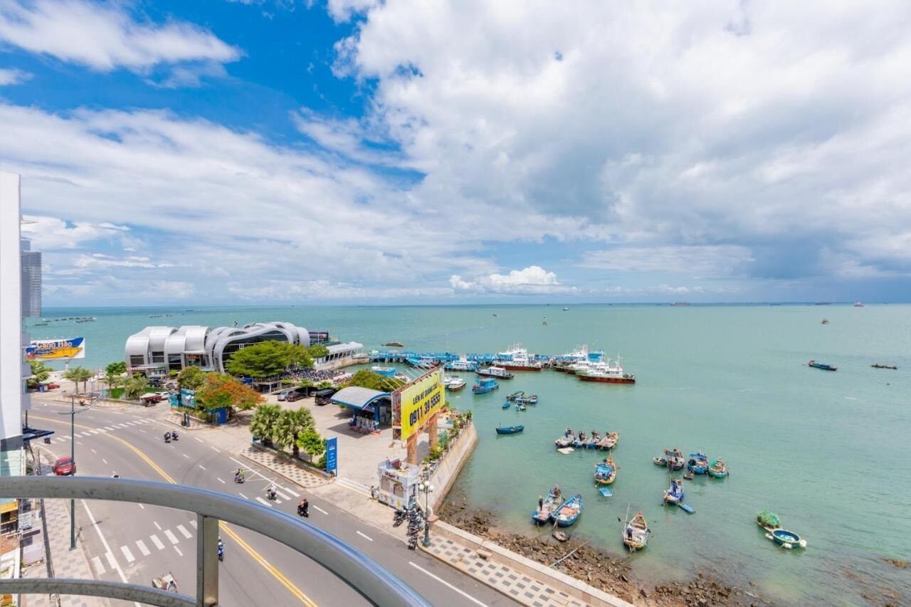 Un paisaje urbano costero con un edificio moderno, un puerto deportivo con barcos y un cielo despejado.