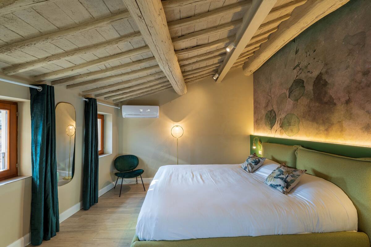 A bedroom with a large bed, a green upholstered headboard, a green chair, a floor lamp, and a wall-mounted air conditioning unit.