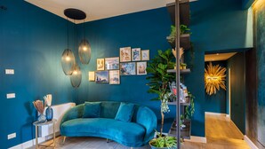 Uma sala de estar moderna com um sofá azul-esverdeado, uma pequena mesa lateral, uma planta alta e uma parede com quadros e um lustre decorativo.