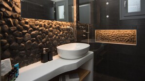 A modern bathroom with a stone wall, a large mirror, a round sink, and a shower area. - Eunoia apartments (Santorini)
