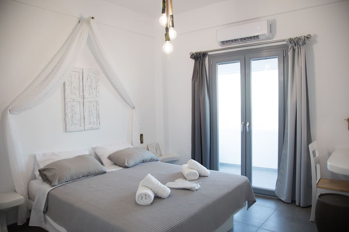 A neatly made bed with a grey bedspread and white pillows, a bedside table with a lamp, and a view of the outside through a sliding glass door.