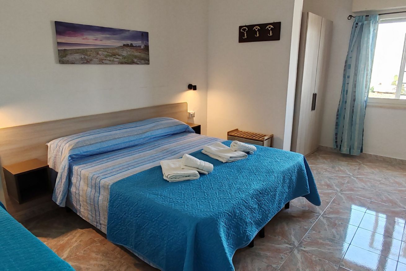 Een slaapkamer met een bed, een nachtkastje, een klein houten krukje, een schilderij aan de muur en een raam met gordijnen.