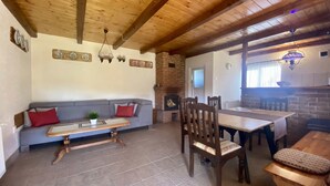 A cozy living room with a wooden ceiling, a brick fireplace, a sofa, a dining table with chairs, and a television.