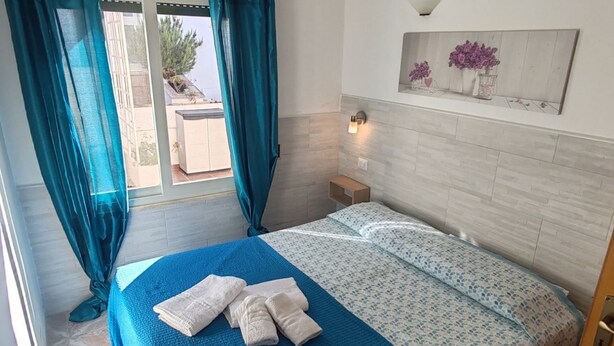 Un dormitorio con cama, mesita de noche, lámpara de pared y ventana con cortinas.