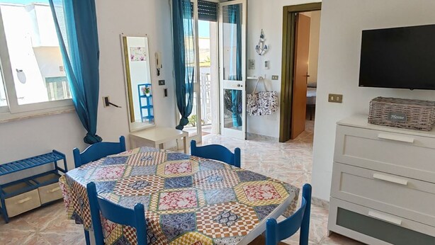 A dining area with a quilted tablecloth, blue chairs, a white cabinet with drawers, and a television on the wall. - Apartment with Veranda in Residence (TORRE SAN GIOVANNI)