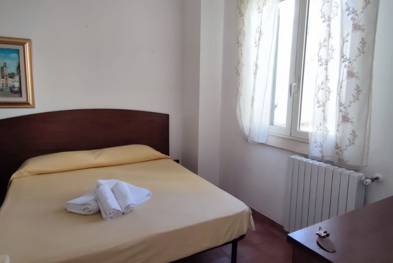 A single bed with a wooden headboard, a yellow bedspread, and two folded white towels. A radiator is visible to the right, and a window with floral curtains is on the wall.