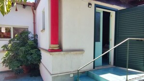 Un escalier menant à une porte à l’extérieur peinte en vert, avec une colonne rouge et un pot de fleurs placé sur le côté de l’escalier.