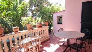 Une terrasse extérieure avec une table ronde et des chaises, entourée de plantes en pot et d’un mur rose.