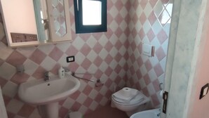 Une salle de bain équipée d’un lavabo, de toilettes et d’un bidet. Les murs sont recouverts de carreaux disposés en motif à carreaux.