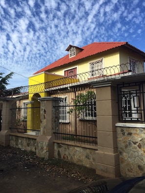 A two-story house with a red roof, yellow walls, and a stone base. It has barred windows and a gated entrance. - Tropical Oasis in Managua (Nindiri)