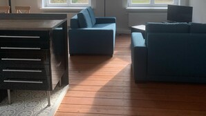 A spacious living room with a wooden floor, two blue sofas, a dining table, and large windows offering a view of trees. - Ferienwohnung am Lindenplatz - Wohnung Weitblick (Baumgarten)