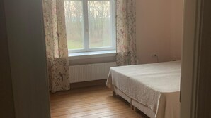 A bedroom with a wooden floor, a bed with a white bedspread, a window with floral curtains, and a hanging light fixture. - Ferienwohnung am Lindenplatz - Wohnung Weitblick (Baumgarten)