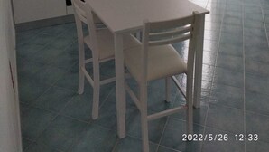 A compact kitchen with white cabinets, a gas stove, a sink, and a refrigerator. There is a dining table with two chairs. The floor is tiled, and the kitchen has a window.
