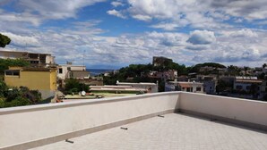 A rooftop terrace with a tiled floor, overlooking a residential area with various buildings and a clear sky with clouds.