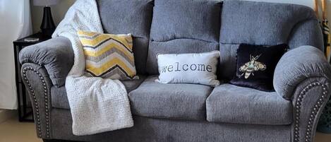 Un sofá gris con tres almohadones: uno bordado con la palabra 'Welcome', otro con un diseño de abeja y un almohadón con estampado de rombos.