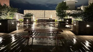 Un'area patio esterna con tavoli e panche in legno, circondata da fioriere e un edificio in mattoni sullo sfondo.