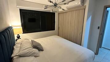 A bedroom with a bed, a nightstand, a wall-mounted air conditioner, a ceiling fan, a wardrobe, and a window.