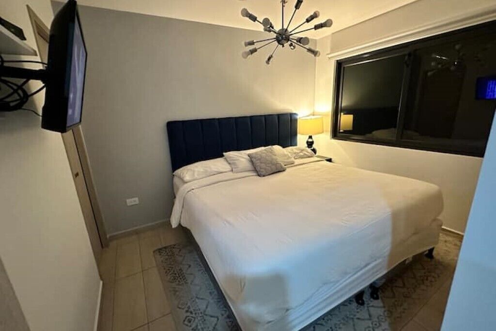 A bedroom with a large bed, a flat-screen TV mounted on the wall, a ceiling light fixture, and a window with a view.