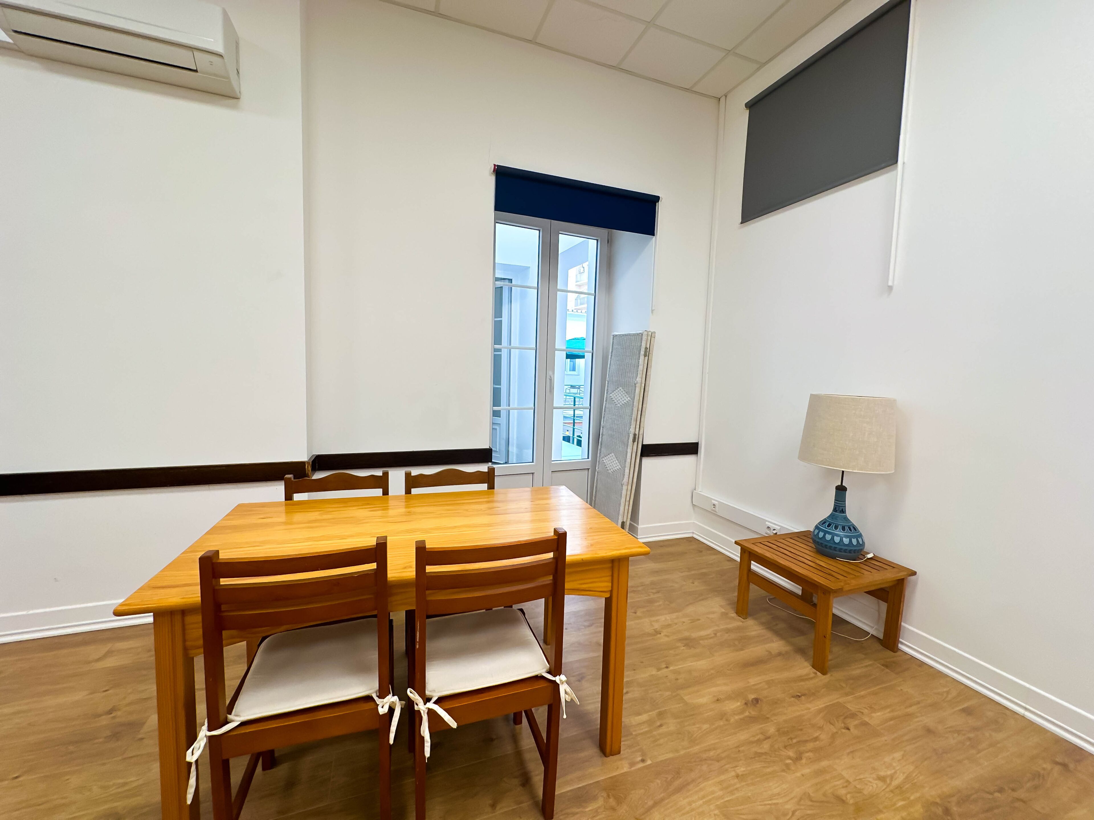 A room with a wooden dining table, chairs, a small wooden side table with a lamp, and a window with a view of buildings.