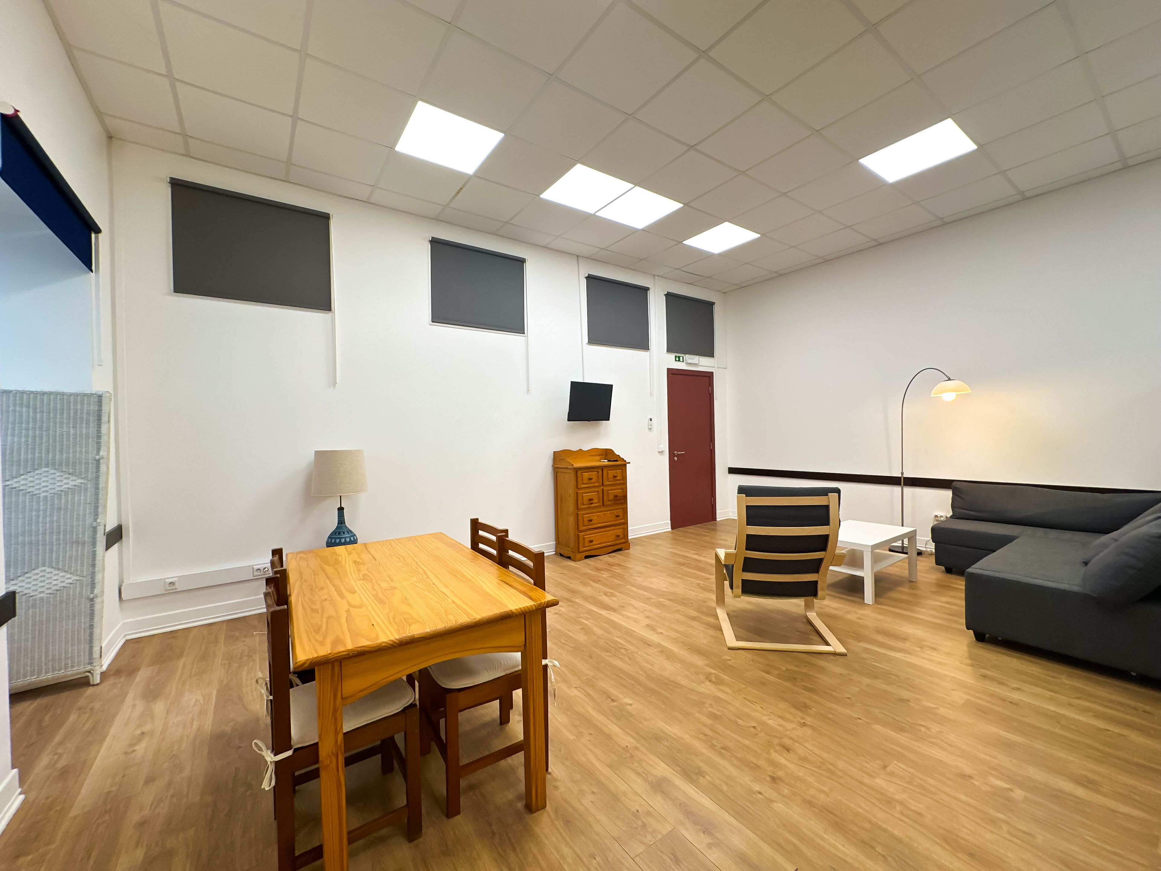 A modern living room with a wooden dining table, a sofa, a lamp, and a TV mounted on the wall.