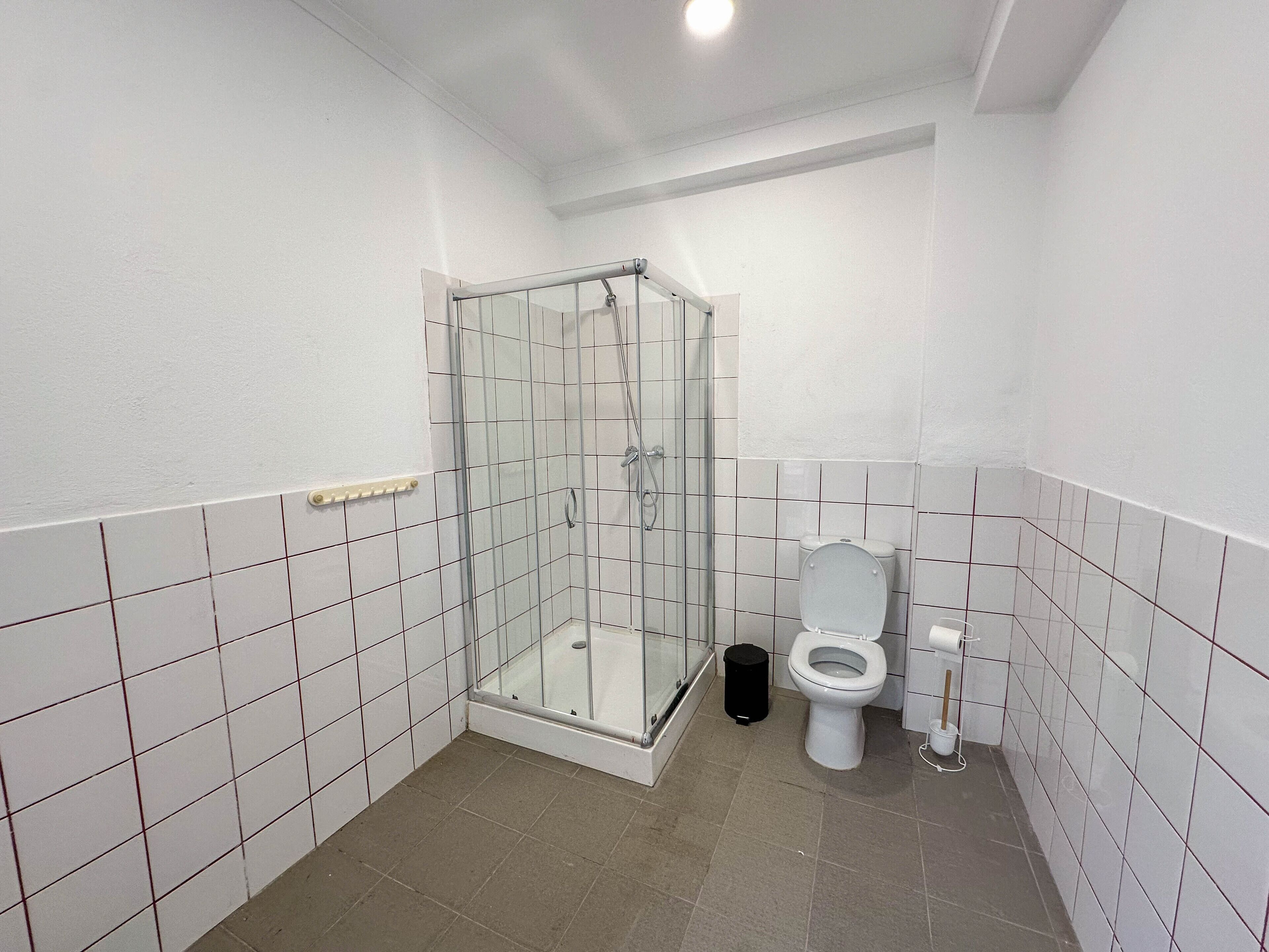 A small, clean bathroom with white tiles, a glass-enclosed shower, a toilet, and a trash bin.