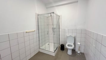 A small, clean bathroom with white tiles, a glass-enclosed shower, a toilet, and a trash bin.