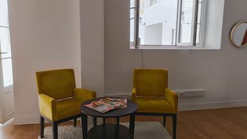 A room with two yellow armchairs, a small round table with magazines, a white shag rug, and a window with a view of a building outside.