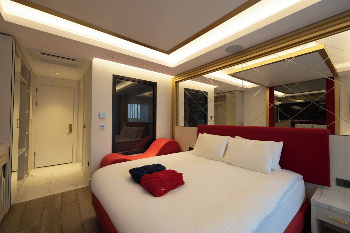 A modern hotel room with a large bed, red headboard, and a mirror above it. There is a door to the left and a small table with a lamp on the right.