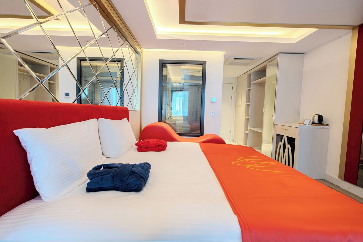 A hotel room with a large bed, a red headboard, a white pillow, a folded towel, and a red blanket. There is a small table with a kettle and a cup, and a mirror on the wall.
