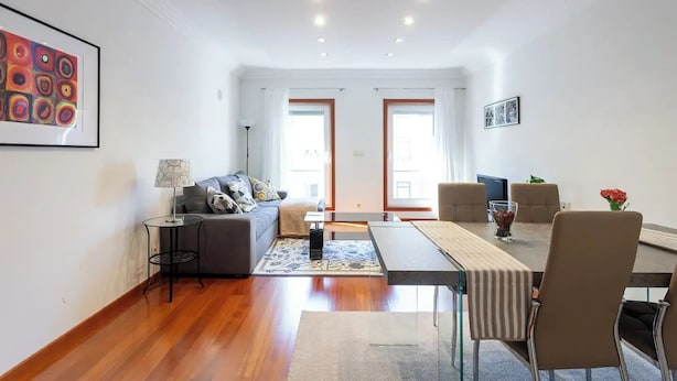 A modern living room with a sofa, coffee table, and dining area. There are two windows with curtains, a lamp, and a framed picture on the wall. - LUXURY LISBOA APARTMENT T2 (Lisbon)