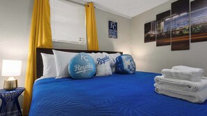 Hotelkamer met een bed met blauw beddengoed, gele gordijnen en kussens met KC Royals branding. Op de muur hangt een foto van het nachtzicht, er staat een lamp en een stapel handdoeken.