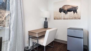 Habitación pequeña con un escritorio de madera, una silla blanca, un refrigerador compacto y un cuadro grande en la pared con un bisonte.