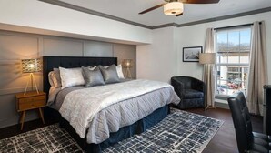 Une chambre avec un grand lit, un ventilateur de plafond, une table de chevet avec une lampe, une fenêtre avec des rideaux et une vue sur l’extérieur.