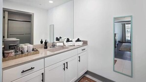 Un baño moderno con un espejo grande, armarios blancos, una encimera con artículos de aseo y una zona de ducha visible.