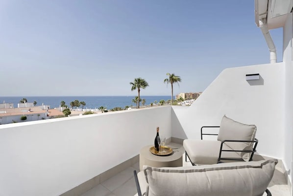 Ein Balkon mit weißem Stuhl, einem kleinen runden Tisch mit Flasche und Glas sowie Blick auf das Meer und Palmen.