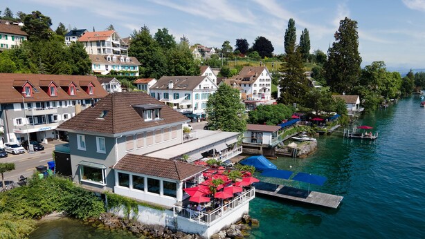 A waterfront property with a large house featuring a balcony with red umbrellas, clear water, and a dock with boats.