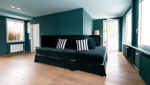A modern bedroom with a dark teal accent wall, a black sofa bed with striped pillows, wooden flooring, and large windows providing natural light.