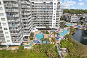A multi-story hotel with balconies, a pool area with multiple pools, and a river nearby.