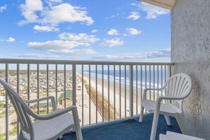 Un balcon avec deux chaises blanches, une vue sur une plage et un parking en arrière-plan.