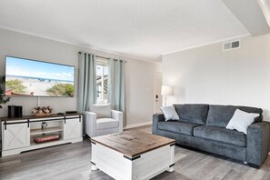 A modern living room with a flat-screen TV, a grey sofa, a white armchair, and a wooden coffee table.