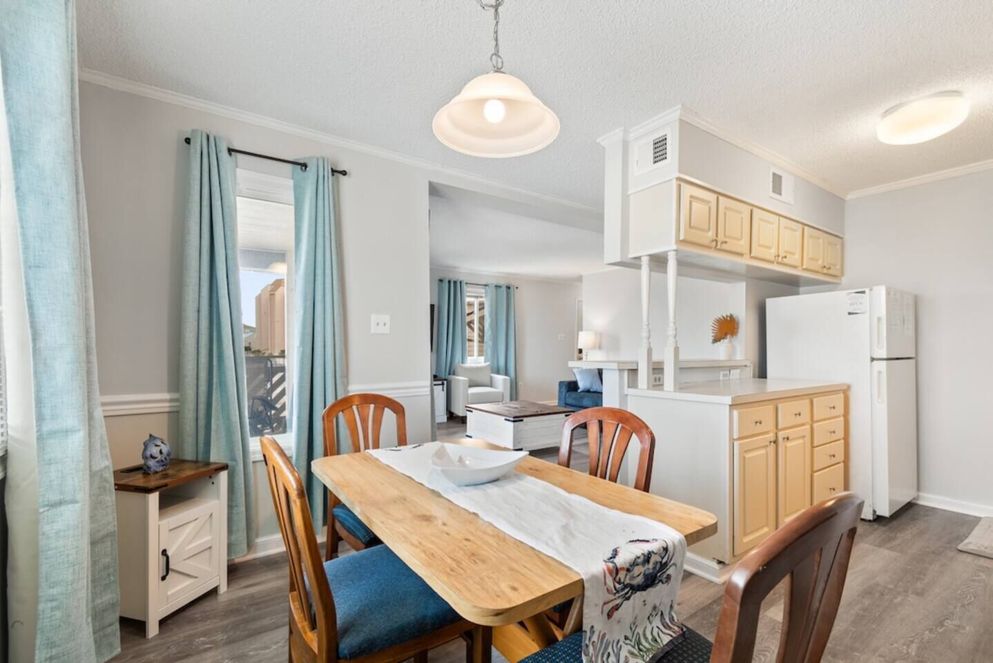 A dining area with a wooden table, chairs, and a white tablecloth. There is a kitchen area with wooden cabinets and a refrigerator.