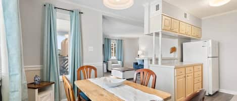 A dining area with a wooden table, chairs, and a white tablecloth. There is a kitchen area with wooden cabinets and a refrigerator.