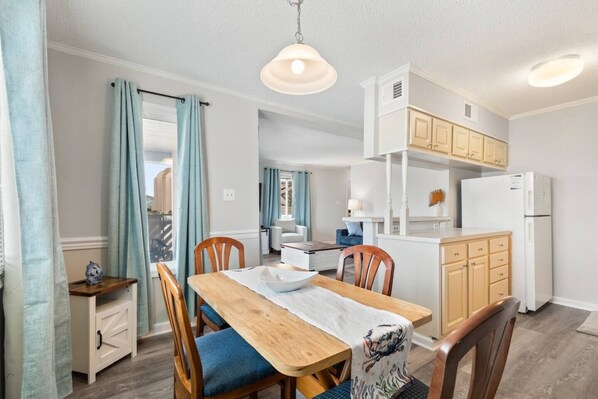 A dining area with a wooden table, chairs, and a white tablecloth. There is a kitchen area with wooden cabinets and a refrigerator.