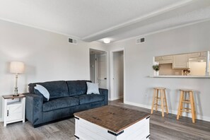 A living room with a dark blue sofa, a wooden coffee table, a lamp, and a kitchen area with stools. - Pools - Near Main St - Beach Steps Away - Waipani (North Myrtle Beach)