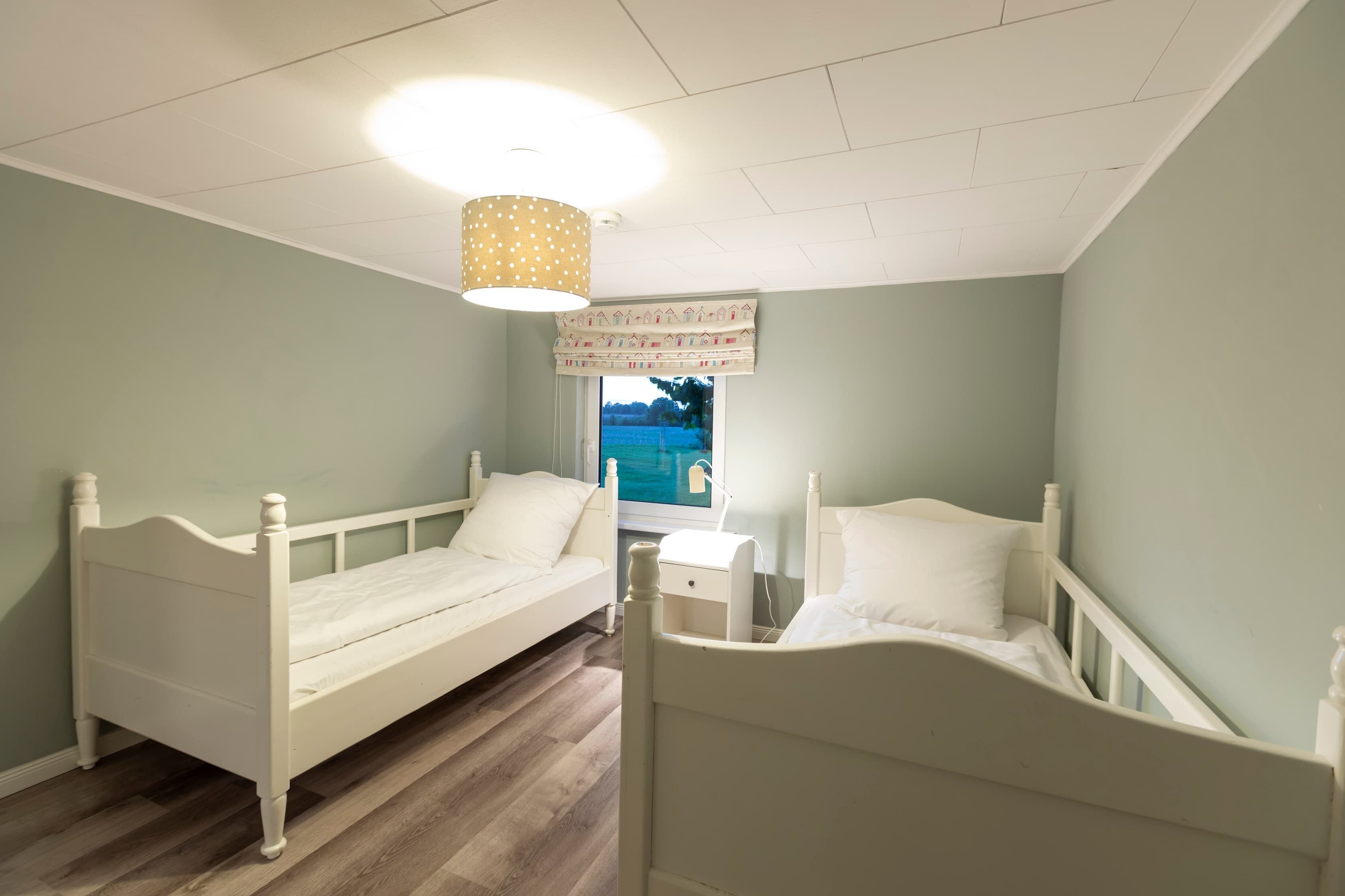 A bedroom with two white beds, a wooden floor, a ceiling light, and a window with a view of greenery.