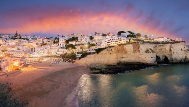 Un pueblo costero con edificios en un acantilado frente al mar, una playa de arena y un cielo al atardecer.