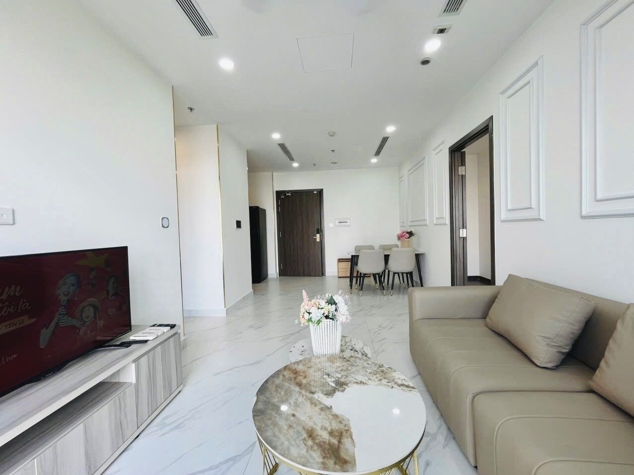 A modern living room with a flat-screen TV, a marble coffee table, a beige sofa, and a dining area in the background.