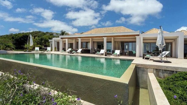 Une piscine avec des chaises longues et des parasols, un bâtiment doté d’un toit en tuiles et un ciel dégagé.