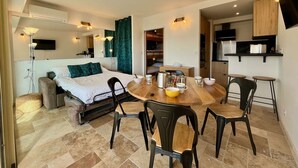 A compact hotel room with a bed, a dining table with chairs, a kitchenette, and a TV mounted on the wall.